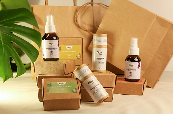 Foto de conjunto exibindo a linha de produtos e embalagens sustentáveis da marca Olga sobre um fundo claro. Dispostas à frente, estão várias caixas de papel kraft para shampoos e condicionadores em barra, dois desodorantes em tubo de papelão e dois frascos de vidro âmbar do Óleo Botânico. Ao fundo, completam o cenário duas sacolas de papel kraft com alças e uma grande folha verde de Monstera no canto esquerdo.