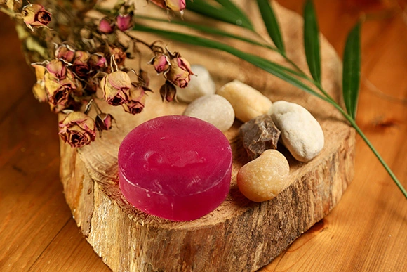 Foto tirada em ângulo superior mostrando um sabonete em barra circular translúcido rosa-violáceo da Olga. O produto está apoiado sobre uma fatia rústica de tronco de madeira maciça, acompanhado de pequenas pedras de rio em tons de bege e cinza, um arranjo de botões de rosas secas e folhas verdes alongadas ao fundo, transmitindo um conceito de autocuidado e conexão com a natureza.